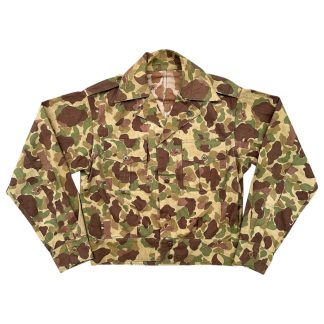 World War II USMC Frogskin camouflage uniform jacket featuring a green and brown disruptive pattern designed for jungle warfare. The jacket includes long sleeves, a button-down front, and two chest pockets with button closures. It has a notched collar and shoulder epaulets, reflecting its military design. The fabric is lightweight and practical for combat in tropical environments.