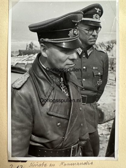 Black-and-white photograph of Field Marshal Erwin Rommel in military uniform, wearing a peaked cap and leather coat, accompanied by another German officer with glasses and an Iron Cross. The background shows a beach scene with debris, possibly during an inspection in Normandy.