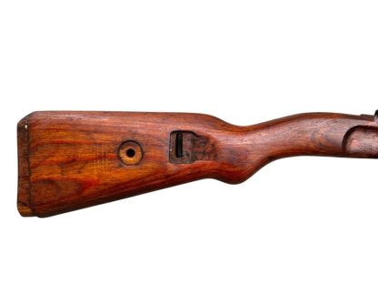 A vintage wooden rifle stock for a German WWII Mauser K98, showcasing a polished reddish-brown finish with visible wood grain texture. The design features a curved grip and cutouts for mounting hardware. The stock appears well-preserved with signs of age and use, such as minor scuffs and wear, adding to its historical authenticity.