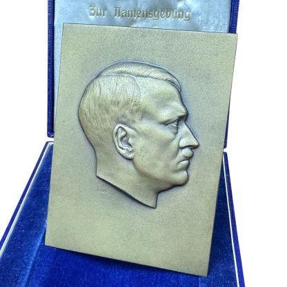 Metal plaque featuring the side profile of Adolf Hitler in relief, presented in a blue velvet box with white interior lining. The text 'Zur Namensgebung' is visible on the inside of the lid. Historical object from the period of the Third Reich.