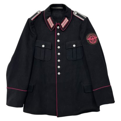 A black German Werkfeuerwehr uniform jacket from World War II, featuring pink piping, metal buttons, shoulder boards, and an emblem with an eagle and swastika on the sleeve. The jacket has two chest pockets and a traditional collar with insignias