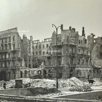 A black and white photograph of war-torn Berlin in 1945, showing the heavily bombed ruins of buildings, including the Kaiserhof Hotel. Rubble covers the streets, and a lone figure walks amidst the devastation. A damaged streetlamp stands in the foreground, emphasizing the destruction caused by World War II.