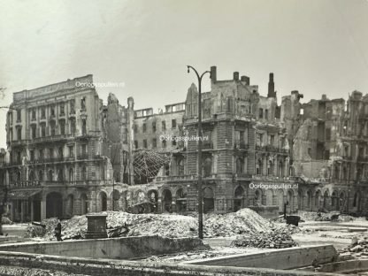 A black and white photograph of war-torn Berlin in 1945, showing the heavily bombed ruins of buildings, including the Kaiserhof Hotel. Rubble covers the streets, and a lone figure walks amidst the devastation. A damaged streetlamp stands in the foreground, emphasizing the destruction caused by World War II.