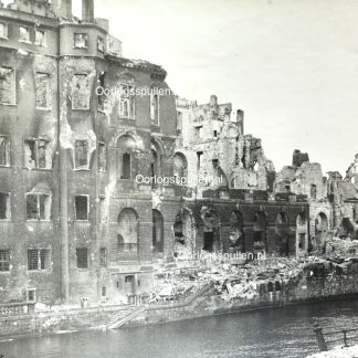 Black and white photograph of heavily bombed buildings in Berlin, Germany, 1945. The structures are partially destroyed, with collapsed walls, shattered windows, and rubble scattered along the riverbank. The devastation is a result of World War II air raids and urban combat.