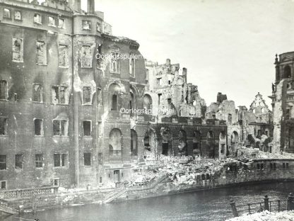 Black and white photograph of heavily bombed buildings in Berlin, Germany, 1945. The structures are partially destroyed, with collapsed walls, shattered windows, and rubble scattered along the riverbank. The devastation is a result of World War II air raids and urban combat.