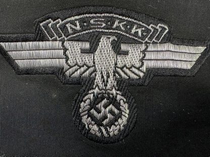 An unissued German NSKK (National Socialist Motor Corps) cap insignia patch from World War II. The black fabric patch features a silver-gray embroidered eagle with outstretched wings, clutching a swastika in its talons, with the letters 'N.S.K.K.' displayed above. The patch appears to be in mint condition, with no visible wear or signs of prior use.