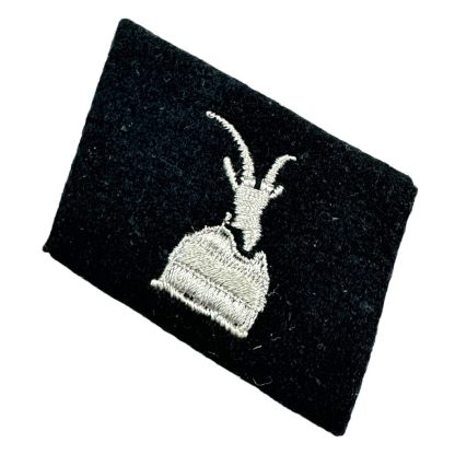 A black fabric collar tab featuring a silver-embroidered mountain goat's head, the insignia of the 21st Waffen Mountain Division of the SS 'Skanderbeg' (Albanian No. 1). The tab is slightly tilted and has a textured surface, representing the WWII-era unit primarily composed of Albanian volunteers.