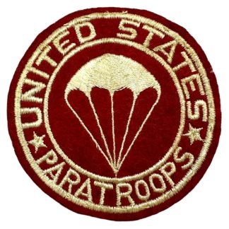 A World War II-era U.S. Airborne Artillery pocket patch. The patch is circular with a red wool background and features embroidered white lettering and design. 'UNITED STATES PARATROOPS' is stitched around the outer edge, separated by two stars. The center showcases a stylized parachute, symbolizing airborne forces. The patch is designed to be worn on the uniform pocket.