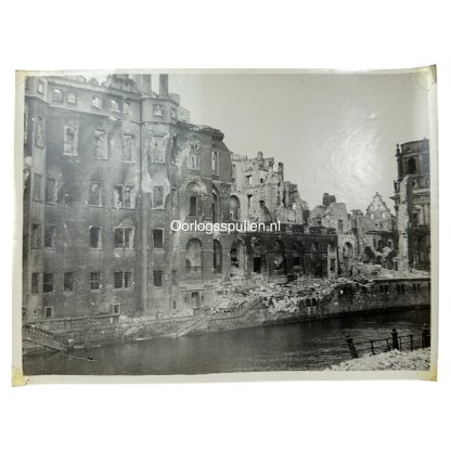 Black and white photograph of heavily bombed buildings in Berlin, Germany, 1945. The structures are partially destroyed, with collapsed walls, shattered windows, and rubble scattered along the riverbank. The devastation is a result of World War II air raids and urban combat.