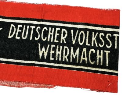 A WWII-era German Volkssturm armband featuring a red fabric background with a black central band, bordered by white stripes. The text 'DEUTSCHER VOLKSSTURM WEHRMACHT' is printed in white, alongside an eagle and swastika emblem. The fabric appears worn with frayed edges.