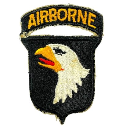 A World War II-era U.S. 101st Airborne Division patch. The patch is black with an embroidered white bald eagle head featuring a yellow beak and red tongue. Above the eagle, a separate curved black tab with 'AIRBORNE' embroidered in yellow thread is attached, though partially torn. The patch shows signs of wear and fraying along the edges.