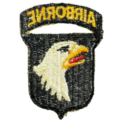 A World War II-era U.S. 101st Airborne Division patch. The patch is black with an embroidered white bald eagle head featuring a yellow beak and red tongue. Above the eagle, a separate curved black tab with 'AIRBORNE' embroidered in yellow thread is attached, though partially torn. The patch shows signs of wear and fraying along the edges.