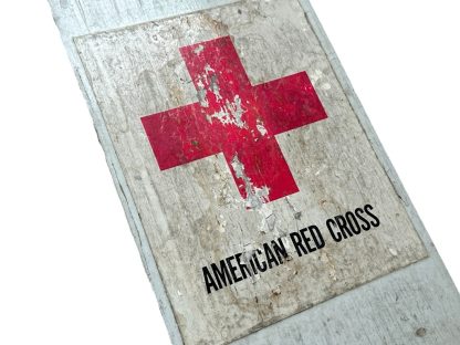 A vintage American Red Cross wooden sign from World War Two, featuring a red cross symbol on a weathered white background with the words 'AMERICAN RED CROSS' printed below. The sign shows significant aging, with chipped paint and worn edges, mounted on a wooden board.