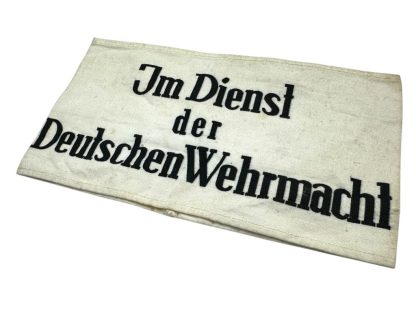 A historical WWII-era German armband made of white fabric with black printed text reading 'Im Dienst der Deutschen Wehrmacht,' meaning 'In the Service of the German Wehrmacht.' The fabric shows signs of aging with minor stains and creases.