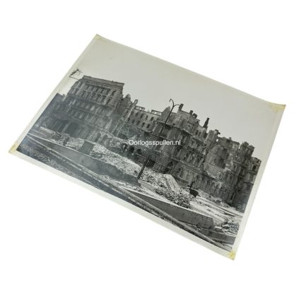 A black and white photograph of war-torn Berlin in 1945, showing the heavily bombed ruins of buildings, including the Kaiserhof Hotel. Rubble covers the streets, and a lone figure walks amidst the devastation. A damaged streetlamp stands in the foreground, emphasizing the destruction caused by World War II.