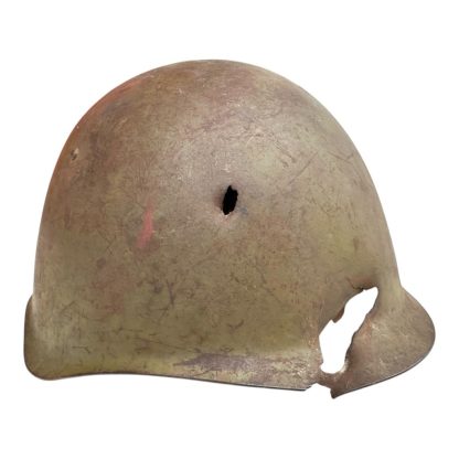 Side view of a battle-damaged WWII Soviet SSh-39 steel helmet. The helmet has a faded greenish-brown patina with scratches and rust. It features a small bullet or shrapnel hole near the upper dome and a large, jagged impact hole along the lower rim, causing significant deformation. The damage suggests it was hit by enemy fire or battlefield debris, reflecting its historical use.