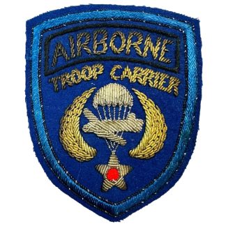 A World War II-era U.S. Airborne Troop Carrier bullion patch, British-made. The patch is shield-shaped with a blue background and features gold bullion embroidery. 'AIRBORNE' is stitched in gold at the top, with 'TROOP CARRIER' below. The central design includes a detailed gold bullion parachute and aircraft flanked by golden wings, with a star at the bottom featuring a red center. The patch has a double blue border, with the outer edge in a lighter blue.