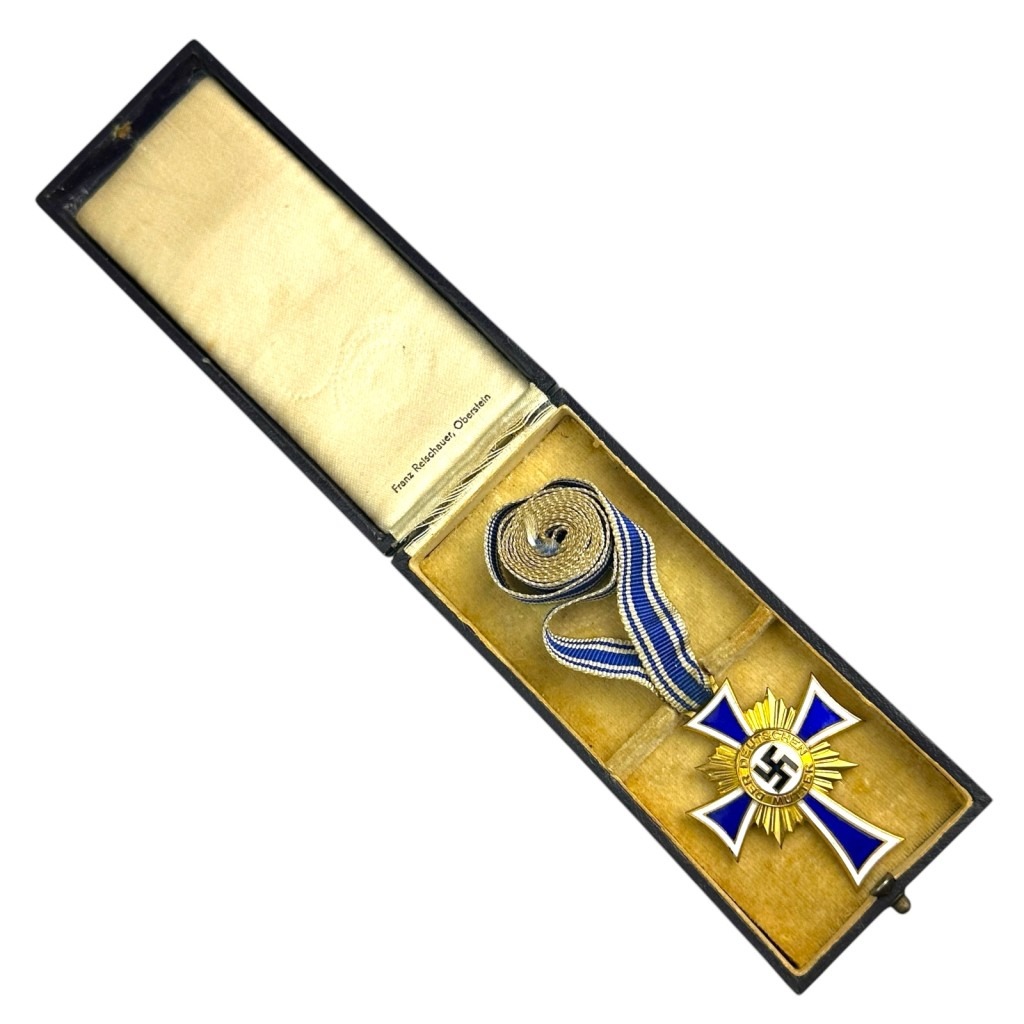 "WWII German Mother’s Cross in Gold (Ehrenkreuz der Deutschen Mutter) in its original presentation case. The gold-enamelled cross with a blue and white ribbon is awarded for mothers with eight or more children. The interior of the case is marked 'Franz Reischauer, Oberstein,' the manufacturer.