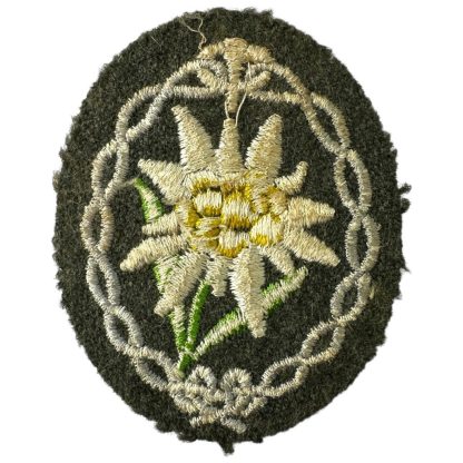 An original WWII German Wehrmacht Gebirgsjäger (mountain troops) arm patch. The oval-shaped patch features an embroidered Edelweiss flower with white petals, a yellow center, and green leaves, surrounded by a decorative silver-grey border, sewn onto a dark green woolen background.