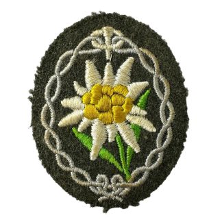 An original WWII German Wehrmacht Gebirgsjäger (mountain troops) arm patch. The oval-shaped patch features an embroidered Edelweiss flower with white petals, a yellow center, and green leaves, surrounded by a decorative silver-grey border, sewn onto a dark green woolen background.