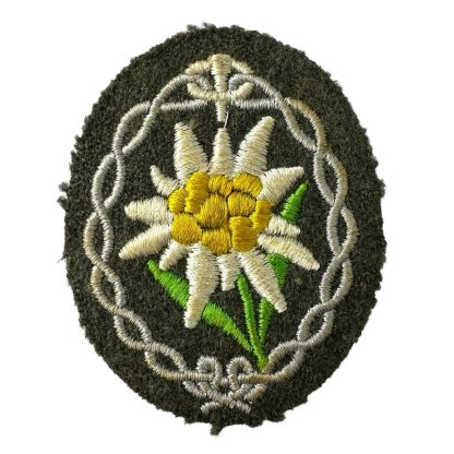 An original WWII German Wehrmacht Gebirgsjäger (mountain troops) arm patch. The oval-shaped patch features an embroidered Edelweiss flower with white petals, a yellow center, and green leaves, surrounded by a decorative silver-grey border, sewn onto a dark green woolen background.