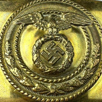 A brass-colored German SA belt buckle featuring an embossed eagle with outstretched wings clutching a wreath that encloses a swastika, surrounded by a decorative circular border."