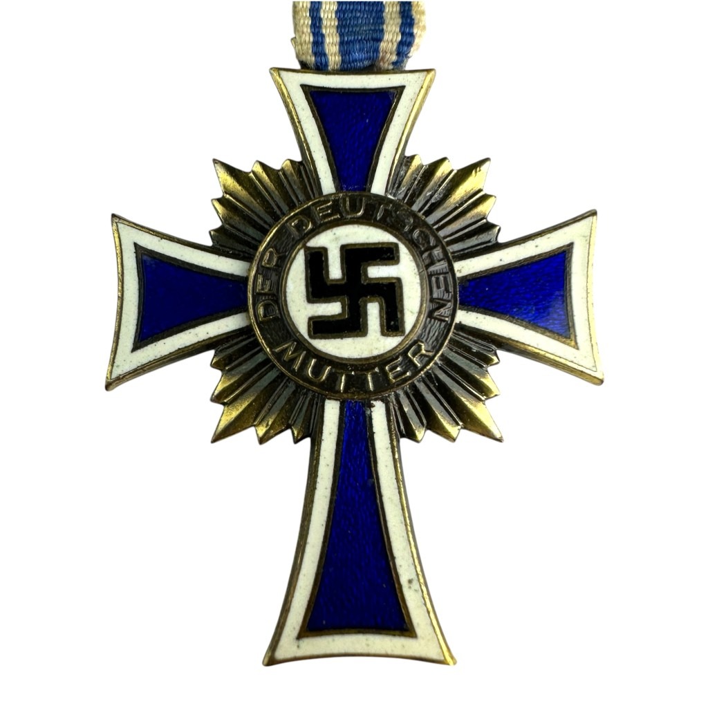A detailed close-up of the German Mother's Cross in Bronze, showing the enameled blue cross, the swastika emblem, and the inscription "DER DEUTSCHEN MUTTER" around the central medallion. The sunburst pattern is visible behind the cross.