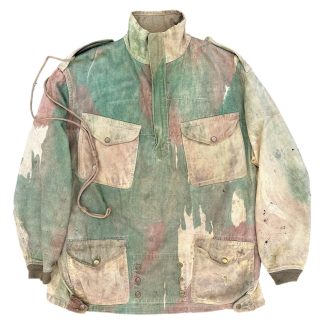 Original WWII British Airborne Denison camouflage smock, 1942, featuring a brushstroke camouflage pattern in green, brown, and tan. The smock has a half-zip front, four front pockets with buttoned flaps, reinforced cuffs, and shoulder epaulets. A distinctive tailpiece with snap buttons is present, designed for securing the smock in place during airborne operations. Signs of wear, fading, and minor damage are visible, indicating authentic wartime use.