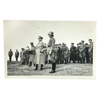 Adolf Hitler, wearing a military trench coat, stands with high-ranking German officers and soldiers during a World War II-era field inspection. The group includes SS and Wehrmacht personnel in uniform and steel helmets, gathered on uneven ground, with the atmosphere suggesting a strategic review or observation.