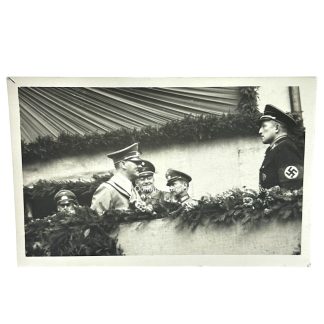 Adolf Hitler stands in a decorated viewing box with several high-ranking Nazi officials, including SS officers, during a public or military event. The scene features draped fabric and floral garlands, suggesting a ceremonial occasion. One officer on the right prominently displays a swastika armband.