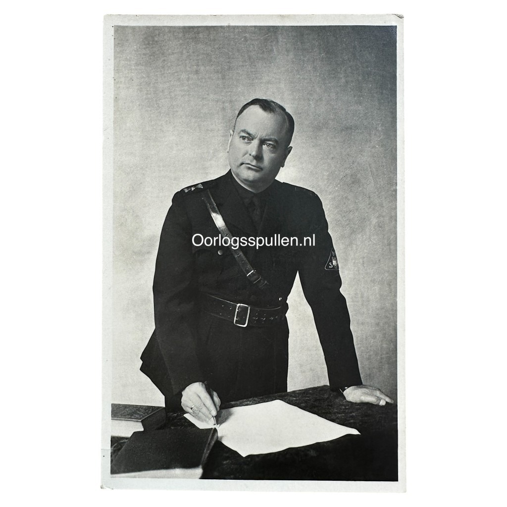 WWII-era Dutch NSB propaganda postcard showing Anton Mussert in full NSB uniform, standing at a desk with pen and paper. Visible insignia and leather cross-strap. Historical collaboration item marked "Oorlogsspullen.nl" – collectible fascist militaria.