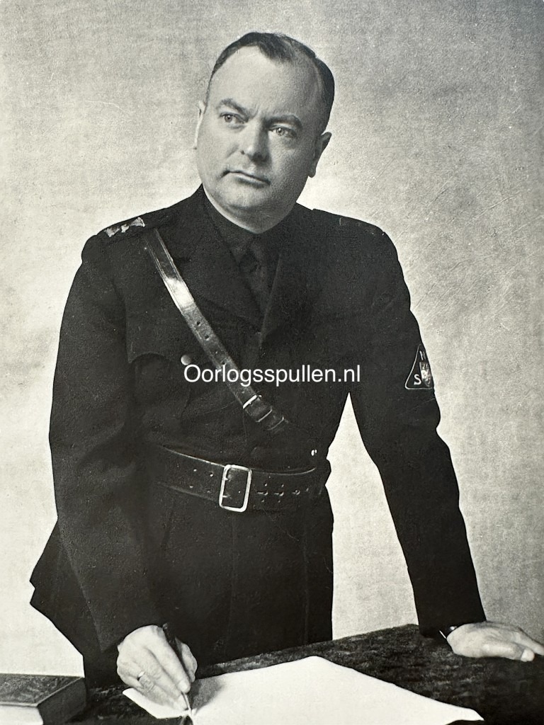 WWII-era Dutch NSB propaganda postcard showing Anton Mussert in full NSB uniform, standing at a desk with pen and paper. Visible insignia and leather cross-strap. Historical collaboration item marked "Oorlogsspullen.nl" – collectible fascist militaria.