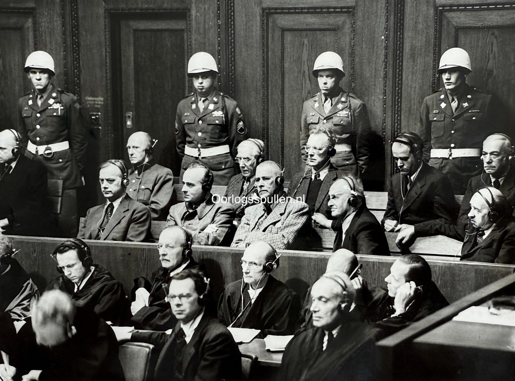 Original British press photo of the Nuremberg Trials in 1946, showing Nazi war criminals in the defendants’ dock wearing headphones for translation, guarded by American military police. Visible are leading figures such as Rosenberg, Frank, Frick, Streicher, Funk, Papen, Speer, Seyss-Inquart, and von Neurath during the International Military Tribunal after World War II.