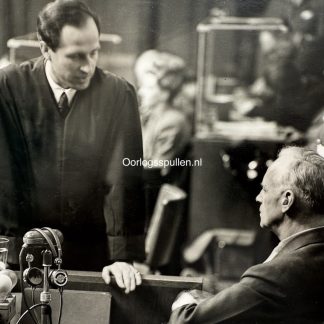 Historic black and white photo of the Nuremberg Trials, showing Nazi foreign minister Joachim von Ribbentrop in the defendant's dock being questioned by a prosecutor, with microphones and military guards present during the International Military Tribunal after World War II.