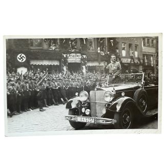Historic black-and-white postcard showing Adolf Hitler standing in an open-top Mercedes-Benz parade car, giving a salute while passing rows of uniformed Nazi Party members performing the Nazi salute. Swastika flags and decorated buildings line the street. Period photograph from the Nazi era.
