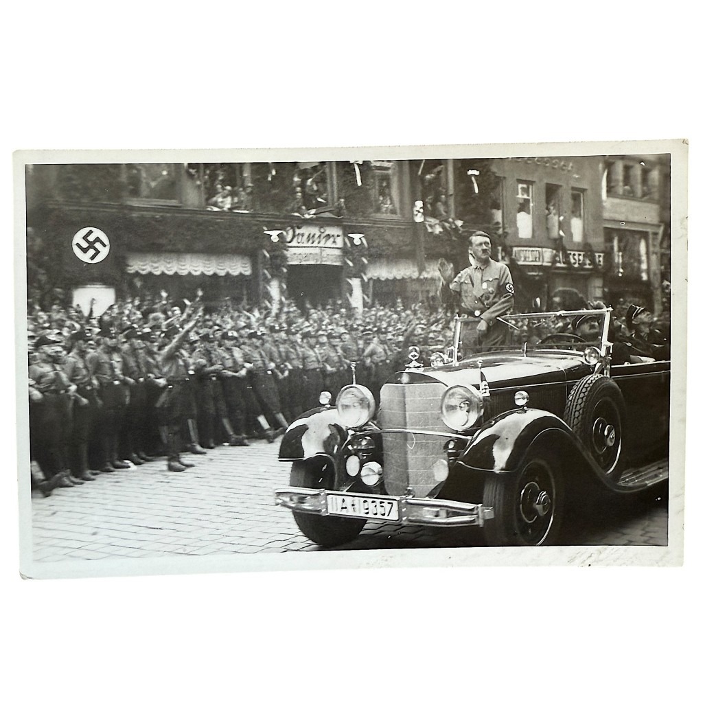 Historic black-and-white postcard showing Adolf Hitler standing in an open-top Mercedes-Benz parade car, giving a salute while passing rows of uniformed Nazi Party members performing the Nazi salute. Swastika flags and decorated buildings line the street. Period photograph from the Nazi era.