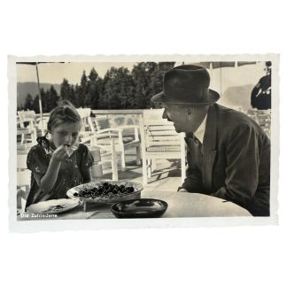 Historic black-and-white postcard with the caption “The Content One.” Adolf Hitler sits at an outdoor table at the Obersalzberg, speaking with a young girl who is eating cherries. Deck chairs and the alpine landscape are visible in the background. Period document from the Nazi era.