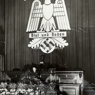 Photo de presse originale de 1937 montrant Hermann Göring prononçant un discours à Berlin devant des représentants de la paysannerie allemande, avec en arrière-plan le grand emblème du Reichsnährstand portant l’aigle, la croix gammée et la devise “Blut und Boden”, ainsi qu’un buste d’Adolf Hitler au-dessus de la tribune.