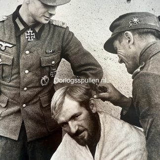 Original WWII photograph showing German Gebirgsjäger soldiers during a break. In the center, a soldier receives a haircut from a comrade, while on the left stands Major Julius Grund, wearing his uniform with the Knight’s Cross of the Iron Cross and other decorations clearly visible. The scene captures a relaxed, human moment among mountain troops.