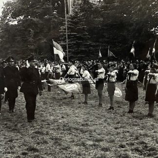 Zwart-witfoto van NSB-leider Anton Mussert tijdens een bezoek aan de Nationale Jeugdstorm (NJS); hij marcheert langs opgestelde jeugdleden die de nationale groet brengen. Op de achtergrond zijn Jeugdstorm-vlaggen en vaantjes (penoenen) zichtbaar. Afmetingen 18 x 13 cm – een interessant origineel stuk voor verzamelaars van Jeugdstorm en NSB-fotomateriaal.