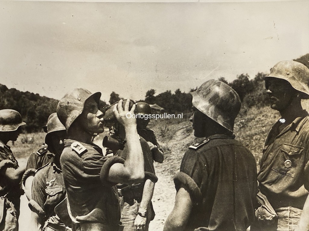 Original WWII German photograph of soldiers from the Panzergrenadier Division Großdeutschland taking a rest in the field, one soldier drinking from a canteen, wearing rare camouflage-painted M42 steel helmets, official German press photo by Kriegsberichter Peiffer dated 14 August 1944, collectible Wehrmacht militaria photo, size 18 x 13 cm.